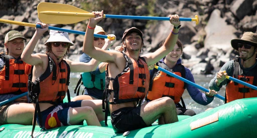 a group of outward bound students sitting in a raft lift their paddles into the air and smile